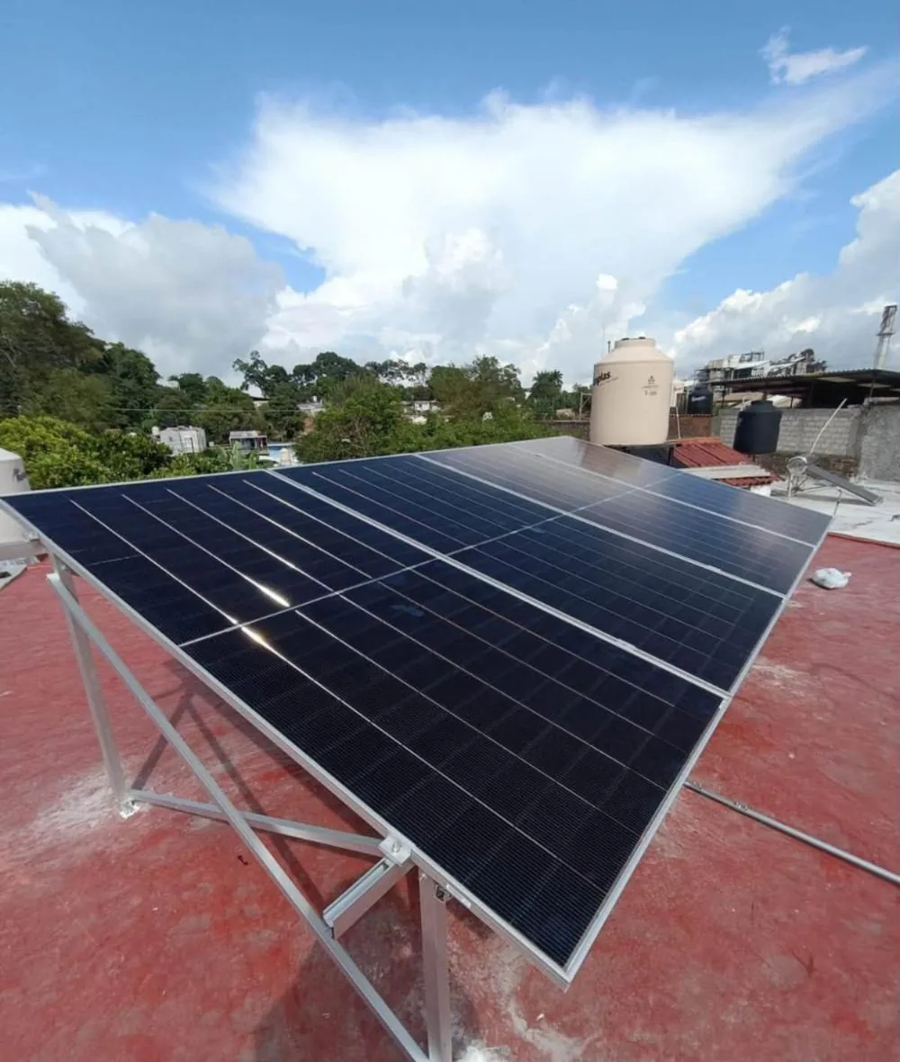 Cuatro paneles solares sobre estructura de aluminio en una azotea roja, con árboles y nubes al fondo